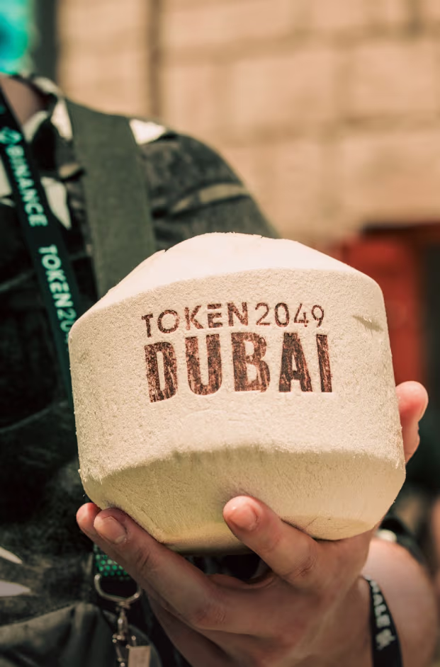 Close-up of a person holding a coconut with the text 'TOKEN 2049 DUBAI' printed on it.