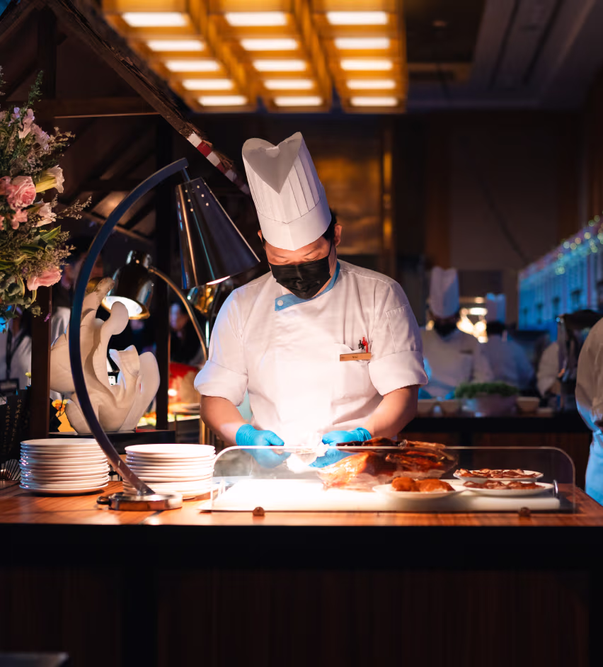 Chef wearing a white uniform, tall hat, black mask, and blue gloves preparing food behind a glass display in a warmly lit kitchen.