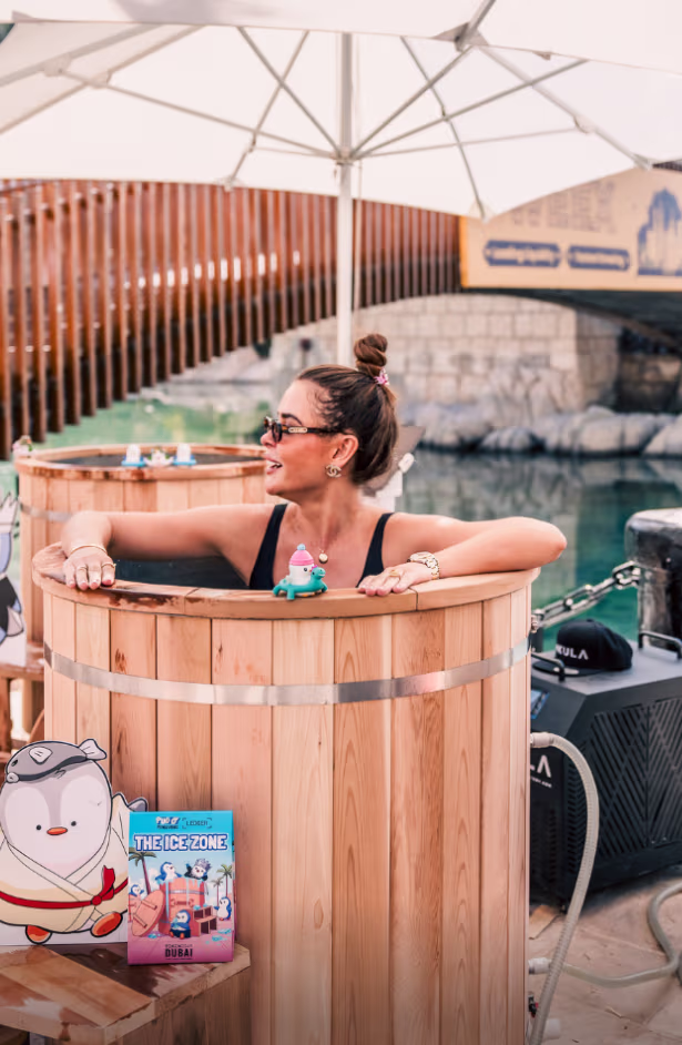 Woman relaxing in a wooden hot tub outdoors near water with a large white umbrella overhead.