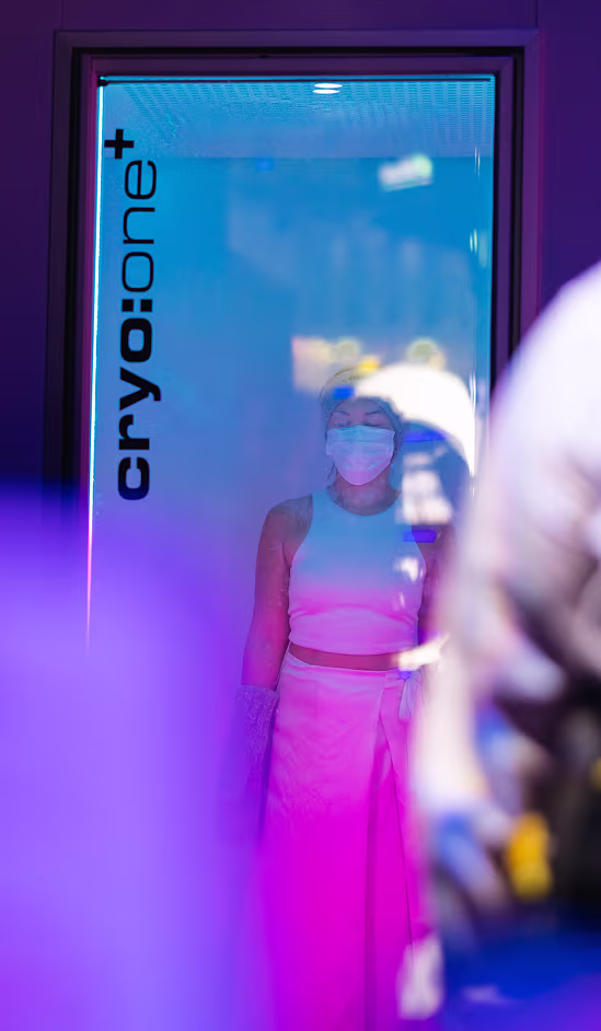 Person wearing a mask standing inside a cryotherapy chamber labeled 'cryo:one+' with blue and purple lighting.