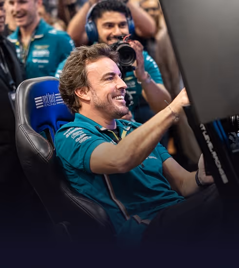 FERNANDO ALONSO in an Aston Martin Racing shirt smiling and using a racing simulator while others watch and photograph him.