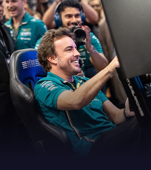 FERNANDO ALONSO in an Aston Martin Racing shirt smiling and using a racing simulator while others watch and photograph him.