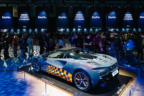 Silver sports car with checkerboard pattern displayed on a platform surrounded by an audience at an indoor event.