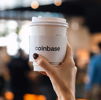 Hand with dark nail polish holding a white takeaway coffee cup with a sleeve labeled 'coinbase' against a blurred background.
