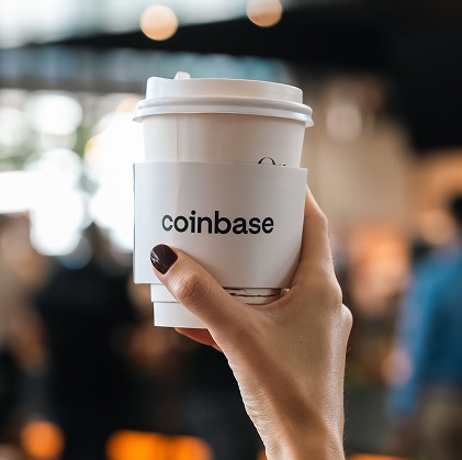 Hand with dark nail polish holding a white takeaway coffee cup with a sleeve labeled 'coinbase' against a blurred background.