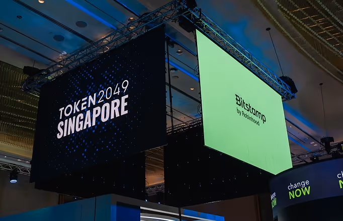 Large hanging screens at an indoor event displaying 'TOKEN2049 SINGAPORE' and 'Bitstamp by Robinhood'.