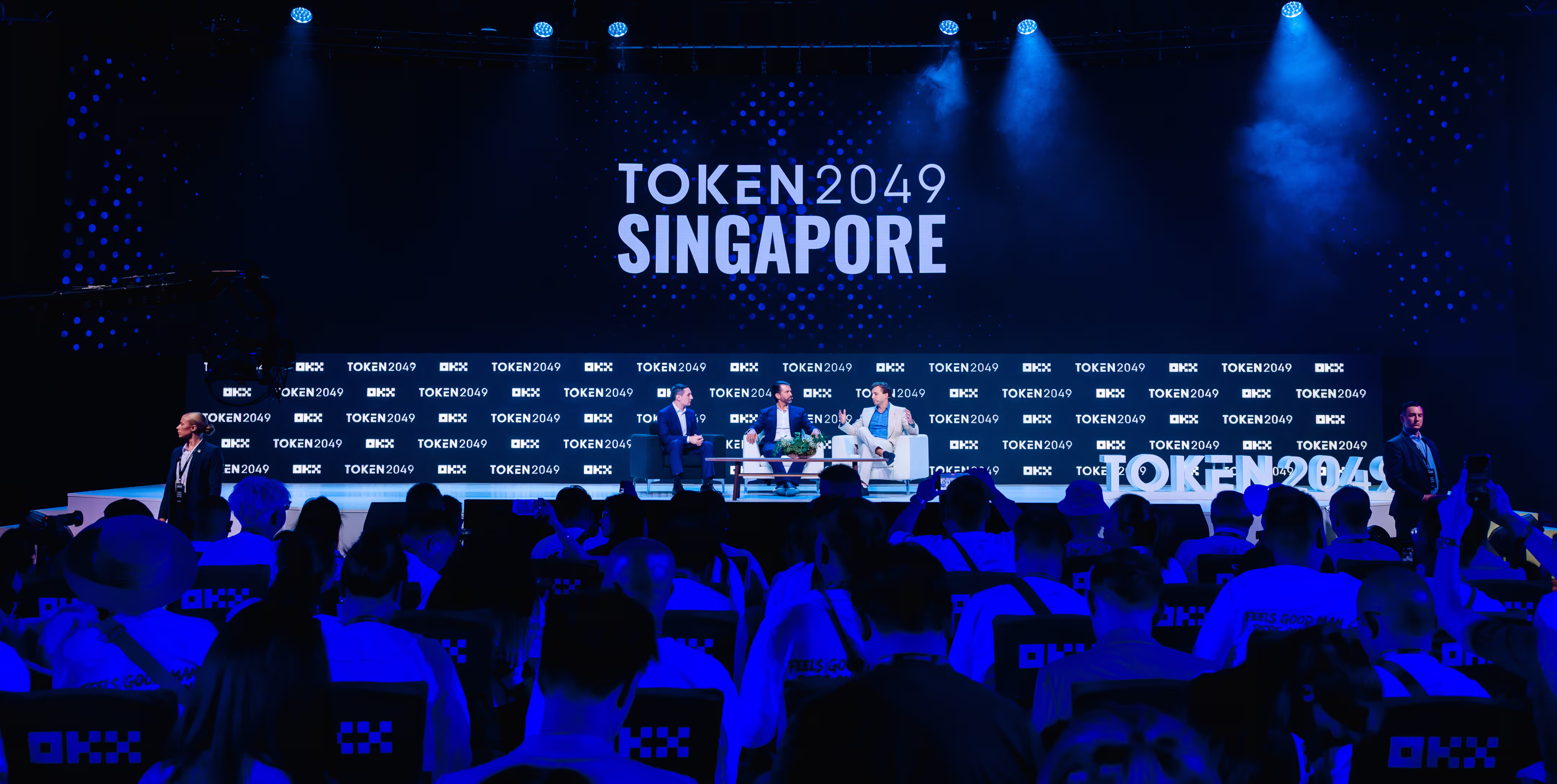 Panel discussion on stage at TOKEN2049 conference in Singapore with an audience watching under blue lighting.