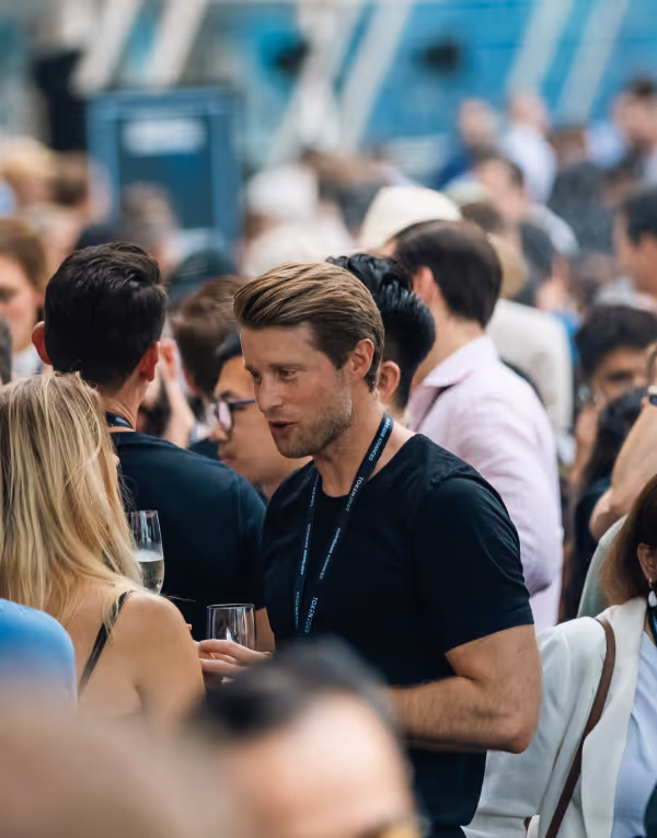 Man in black shirt holding a glass and talking to a blonde woman at a crowded social event.