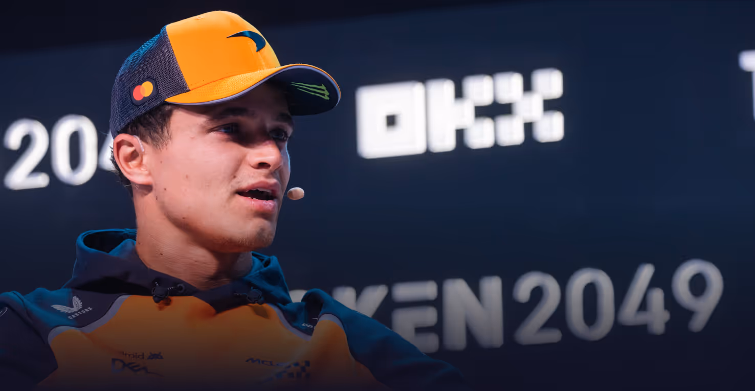 Man in orange and black McLaren racing team jacket and cap speaking with a headset microphone.