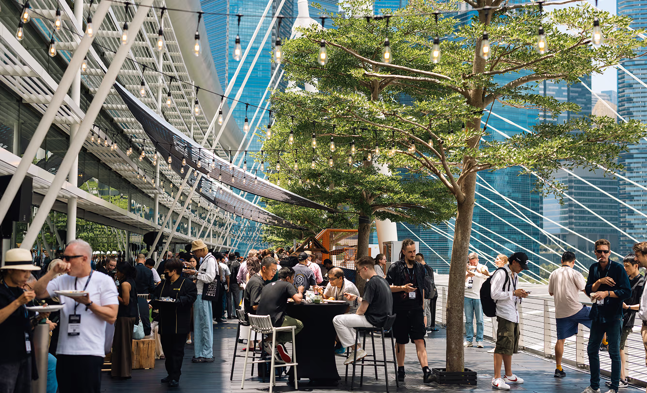 People socializing and eating at an outdoor event under string lights on a modern terrace with trees and skyscrapers in the background.
