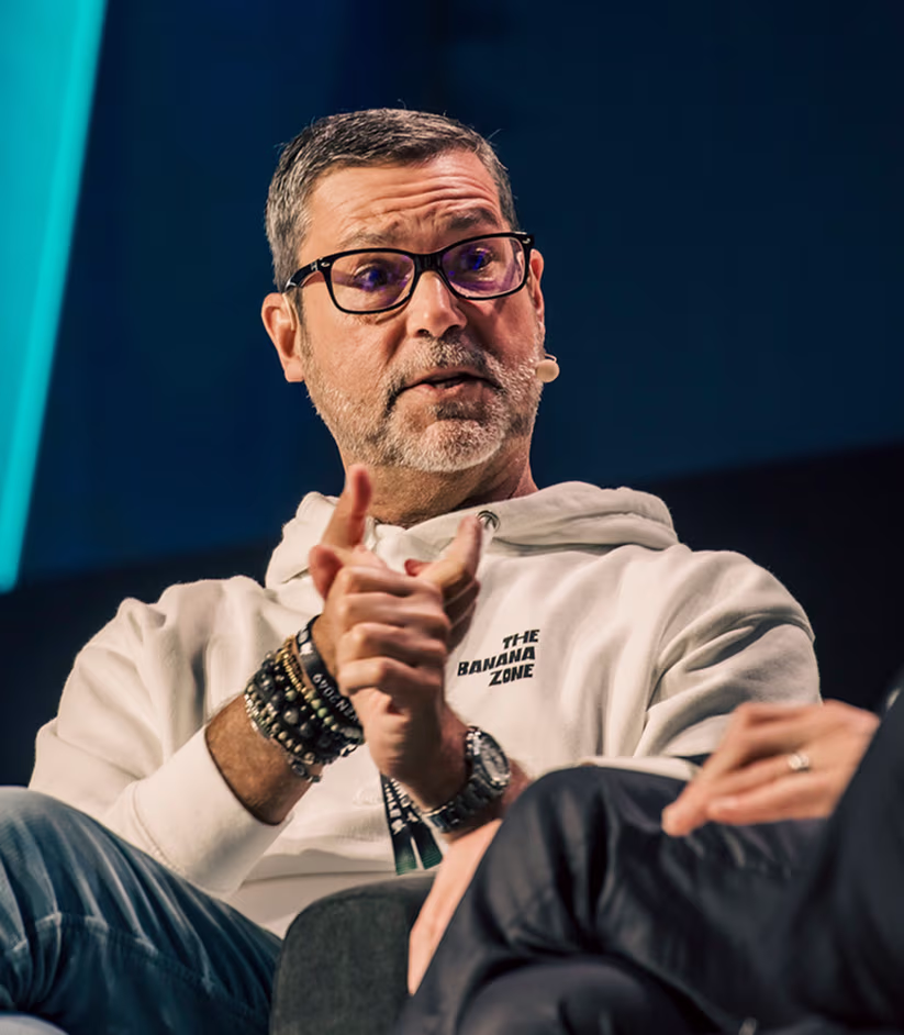 Man with glasses wearing a white hoodie labeled 'The Banana Zone' speaking with hands clasped during a discussion.