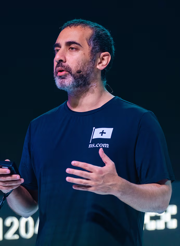 Man with short dark hair and beard speaking on stage wearing a black t-shirt with a flag logo and holding a remote control.