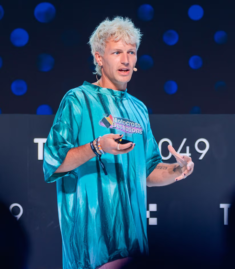 Person with blonde hair speaking on stage wearing a blue Motocross Des Nations shirt and a headset microphone.