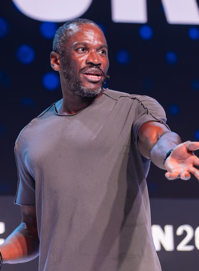 Man speaking on stage with a microphone headset, wearing a gray t-shirt and gesturing with his hand.