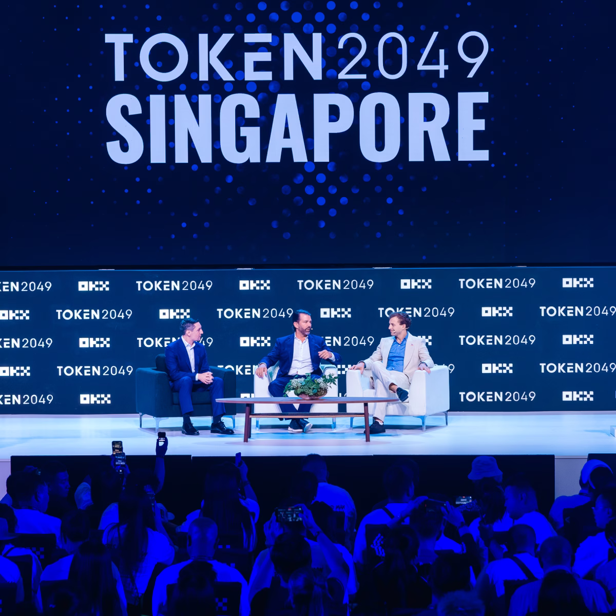 Panel discussion on the TOKEN2049 Singapore main stage with three speakers seated in front of a large screen and audience