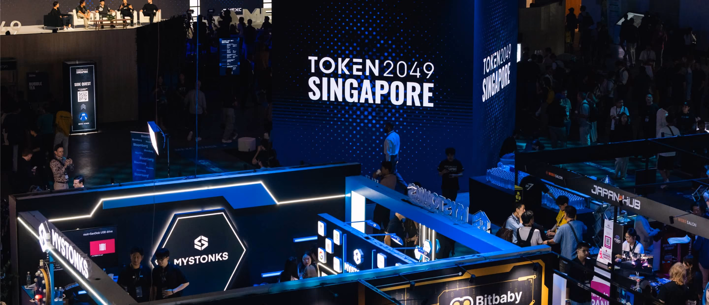 Conference scene with booths and a large display reading TOKEN 2049 SINGAPORE and attendees interacting.