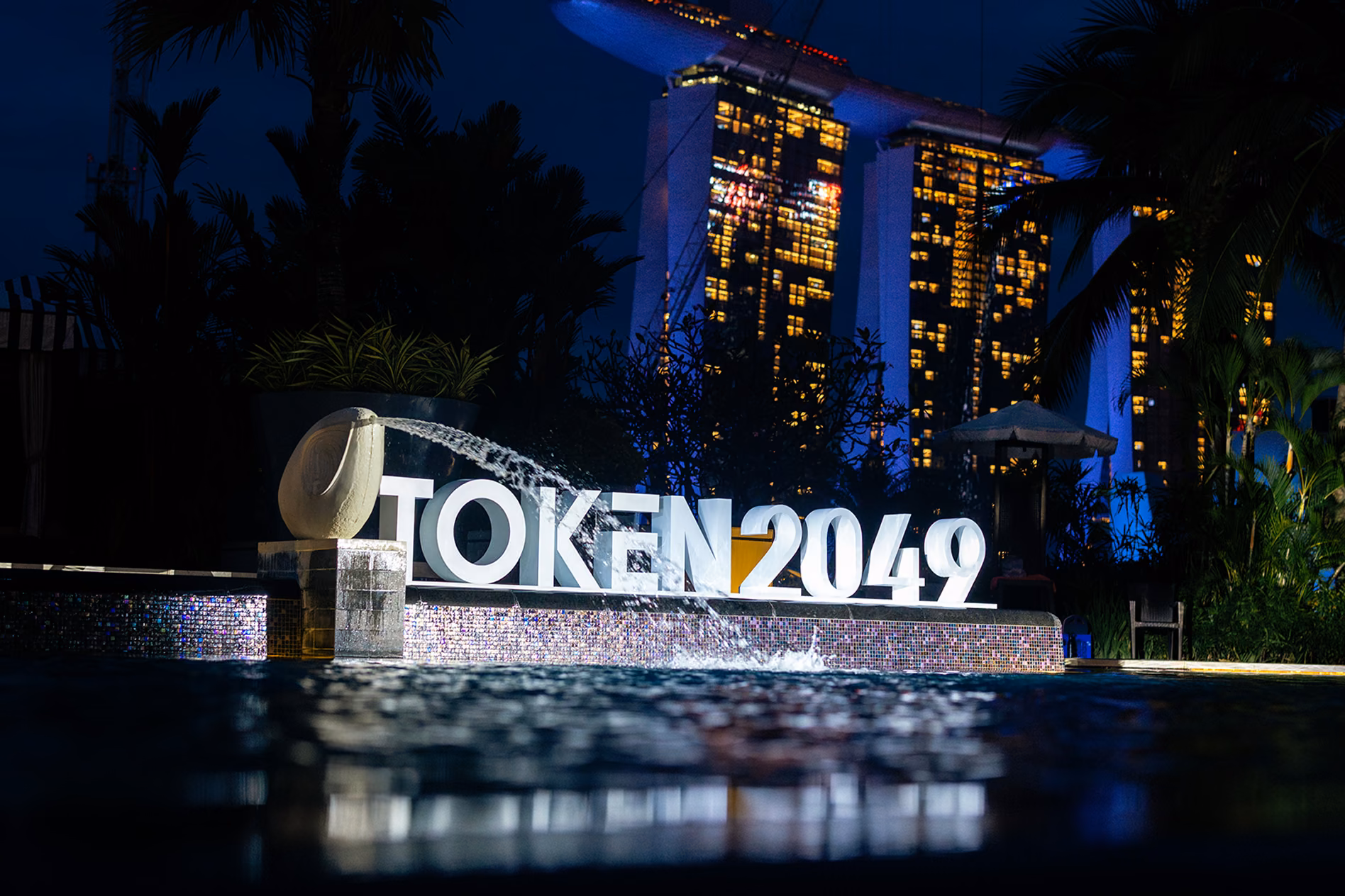 Night view of illuminated 'TOKEN 2049' sign with water fountain in front, reflecting on water surface, and tall buildings with lights in the background.