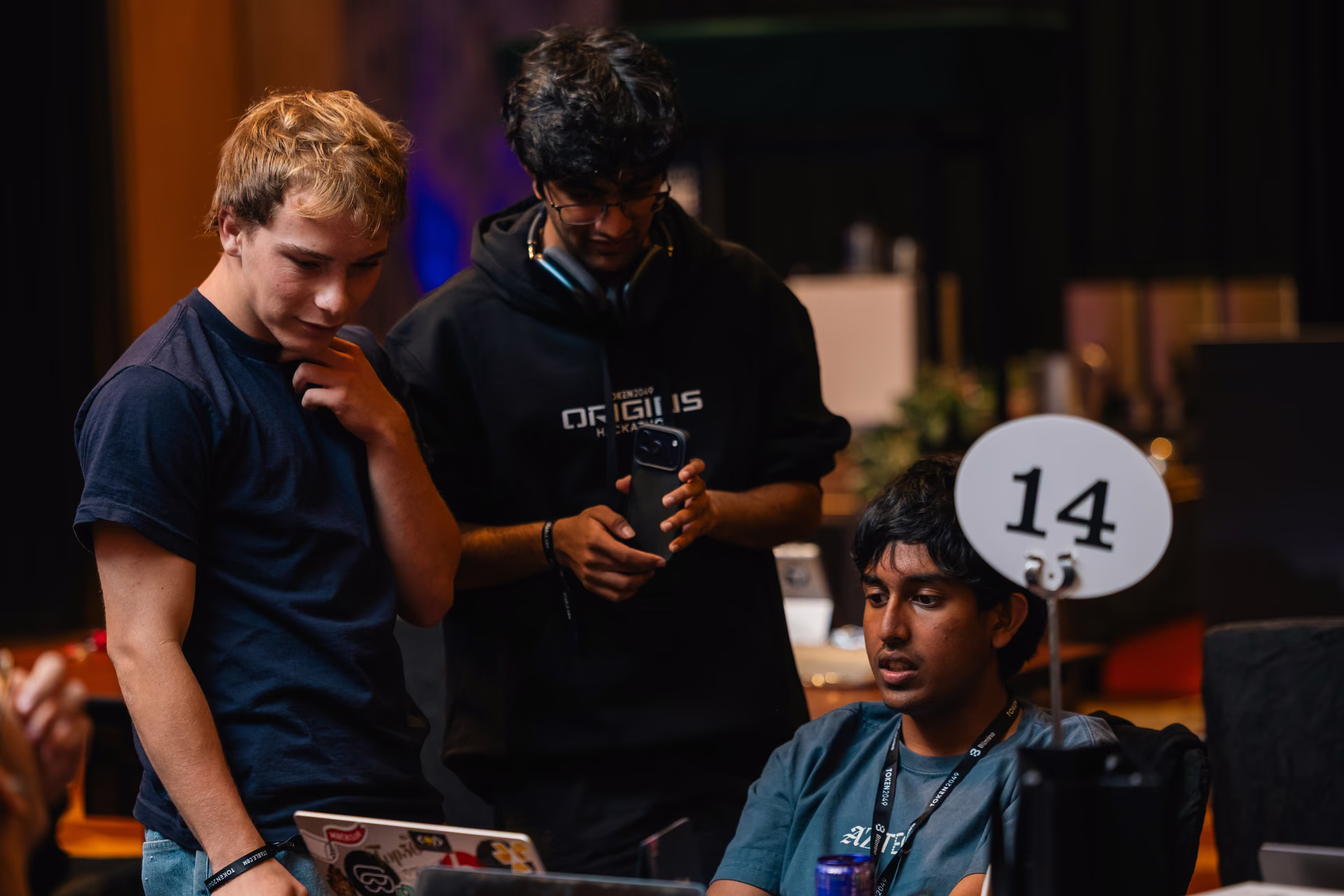 Three young men standing and sitting around a laptop, engaged in discussion, with a table number 14 sign visible.