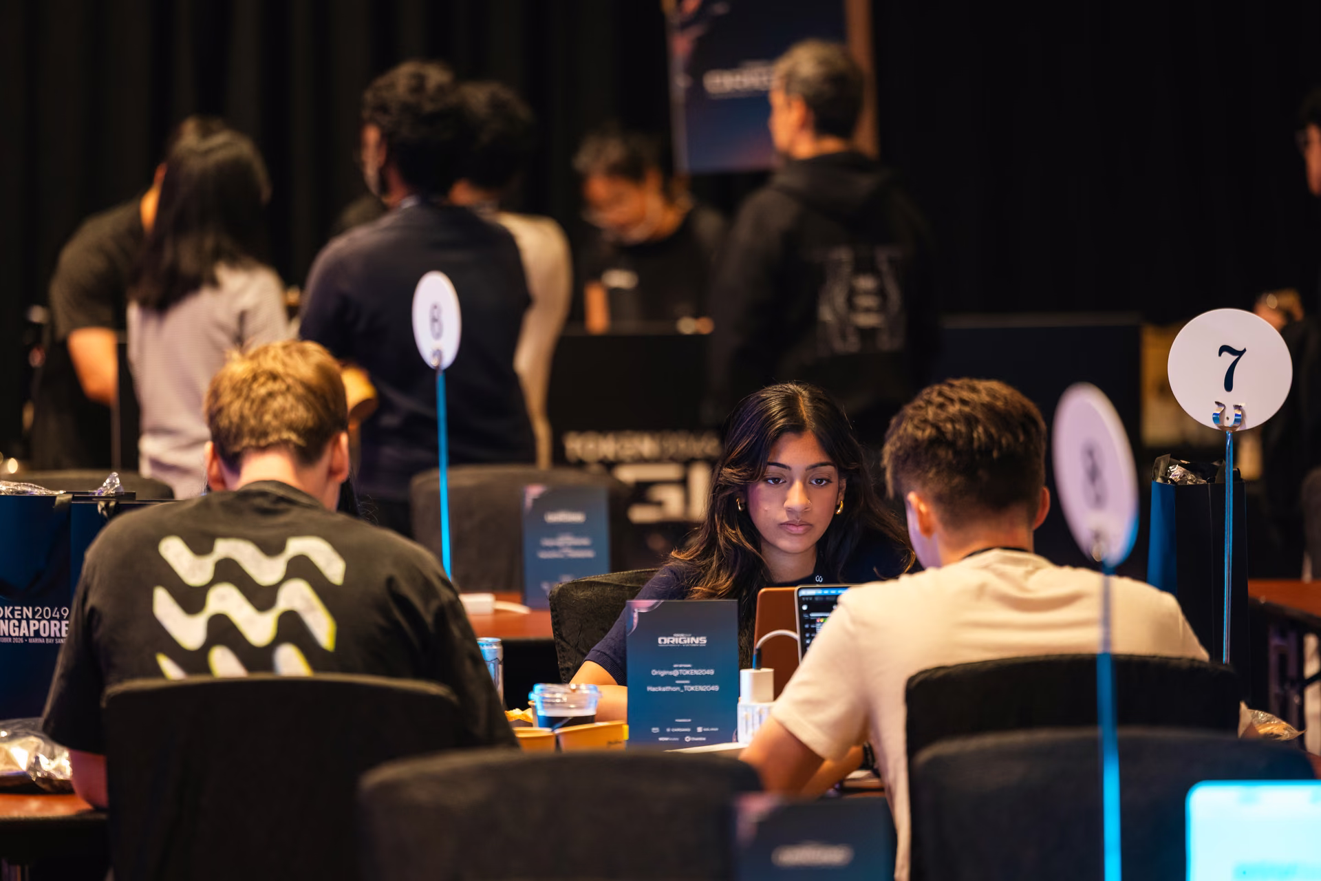 Young woman sitting at a table between two young men, focused on a laptop screen, in a busy indoor event space with other people and numbered table signs visible.