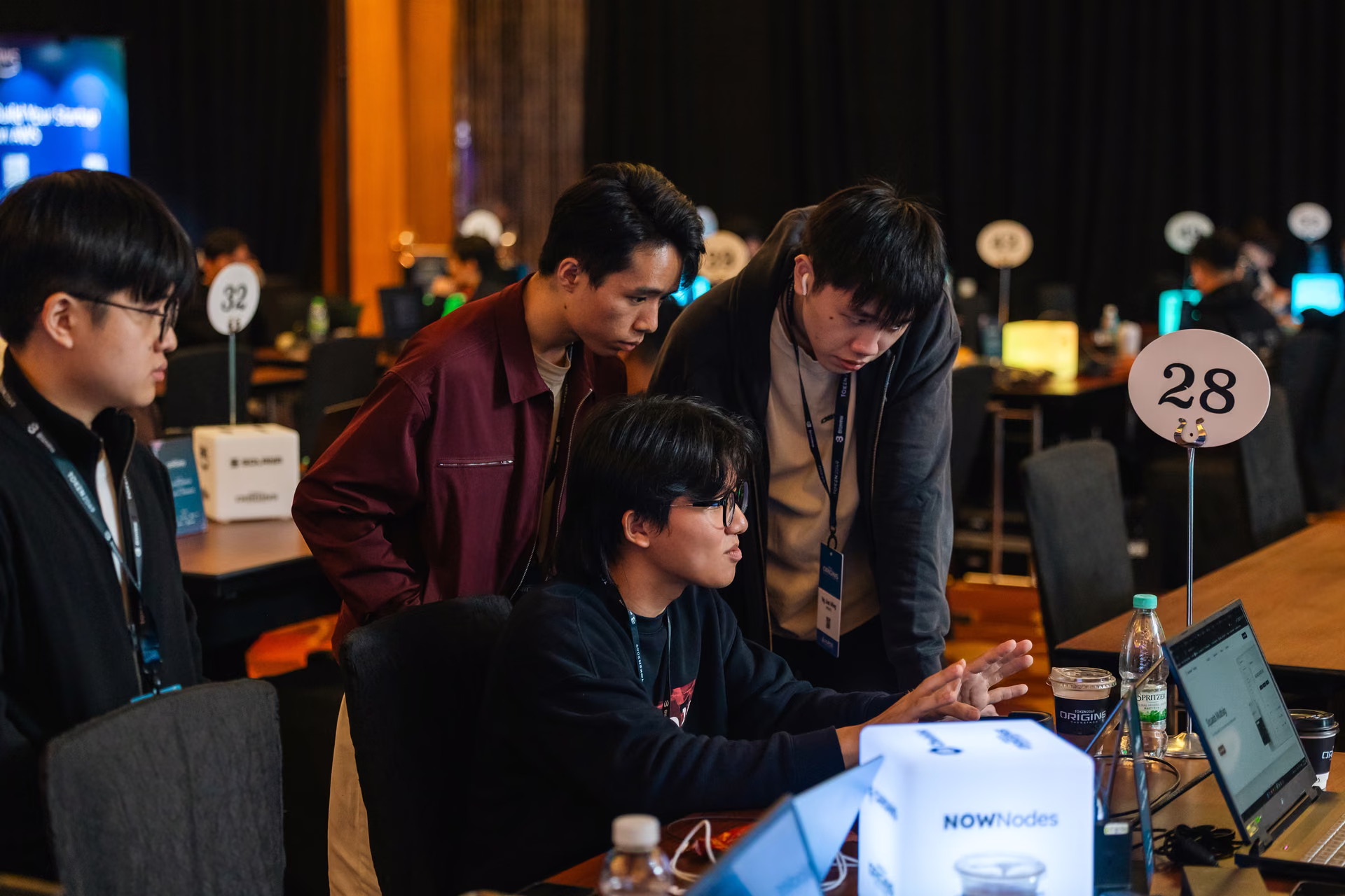 Four young men collaborating in a dimly lit room with computers, focusing on one laptop at table 28, with tech event badges around their necks.