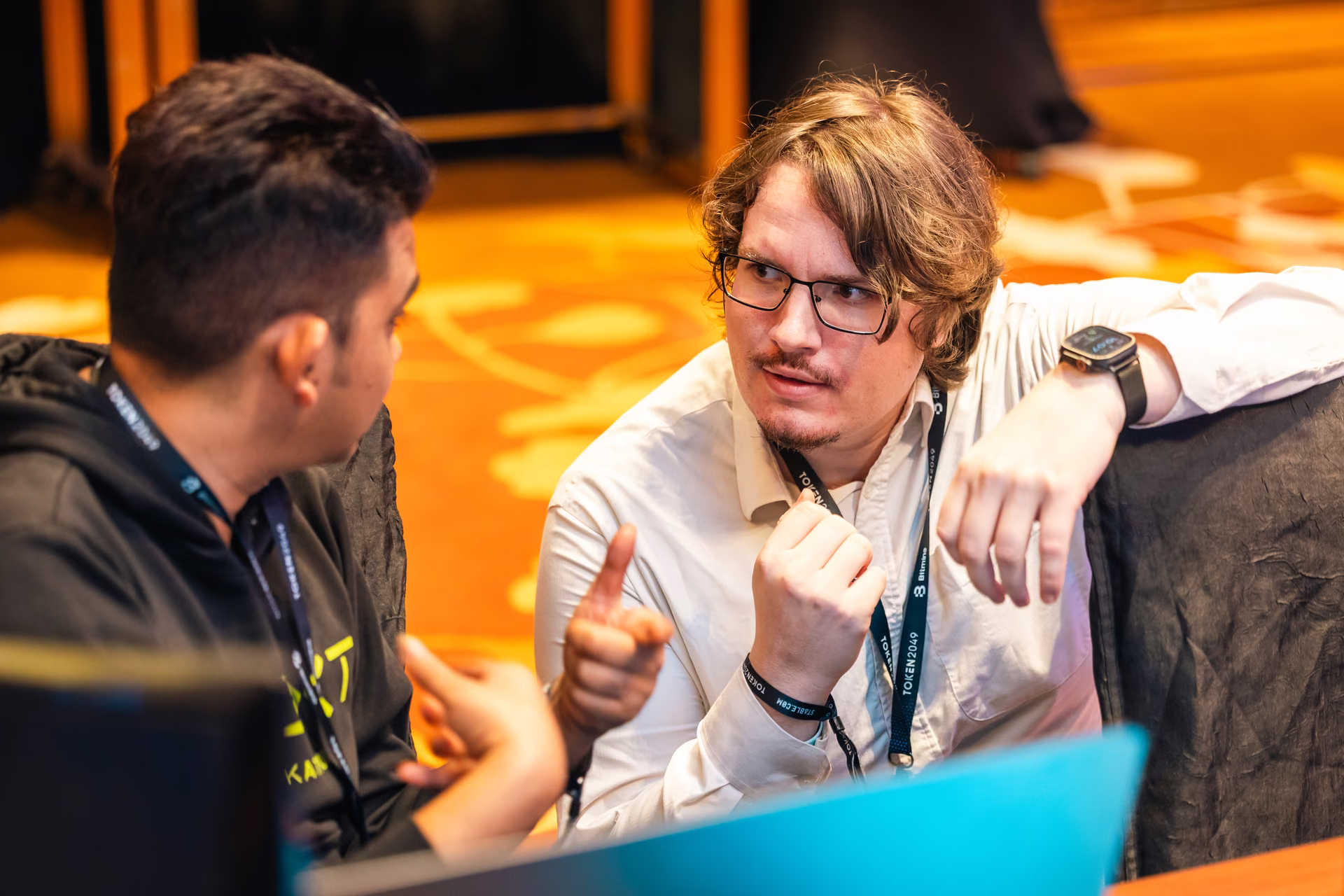 Two men engaged in a discussion, one wearing glasses and a white shirt listens intently while the other gestures with his hands, sitting close together in an indoor setting with warm lighting.
