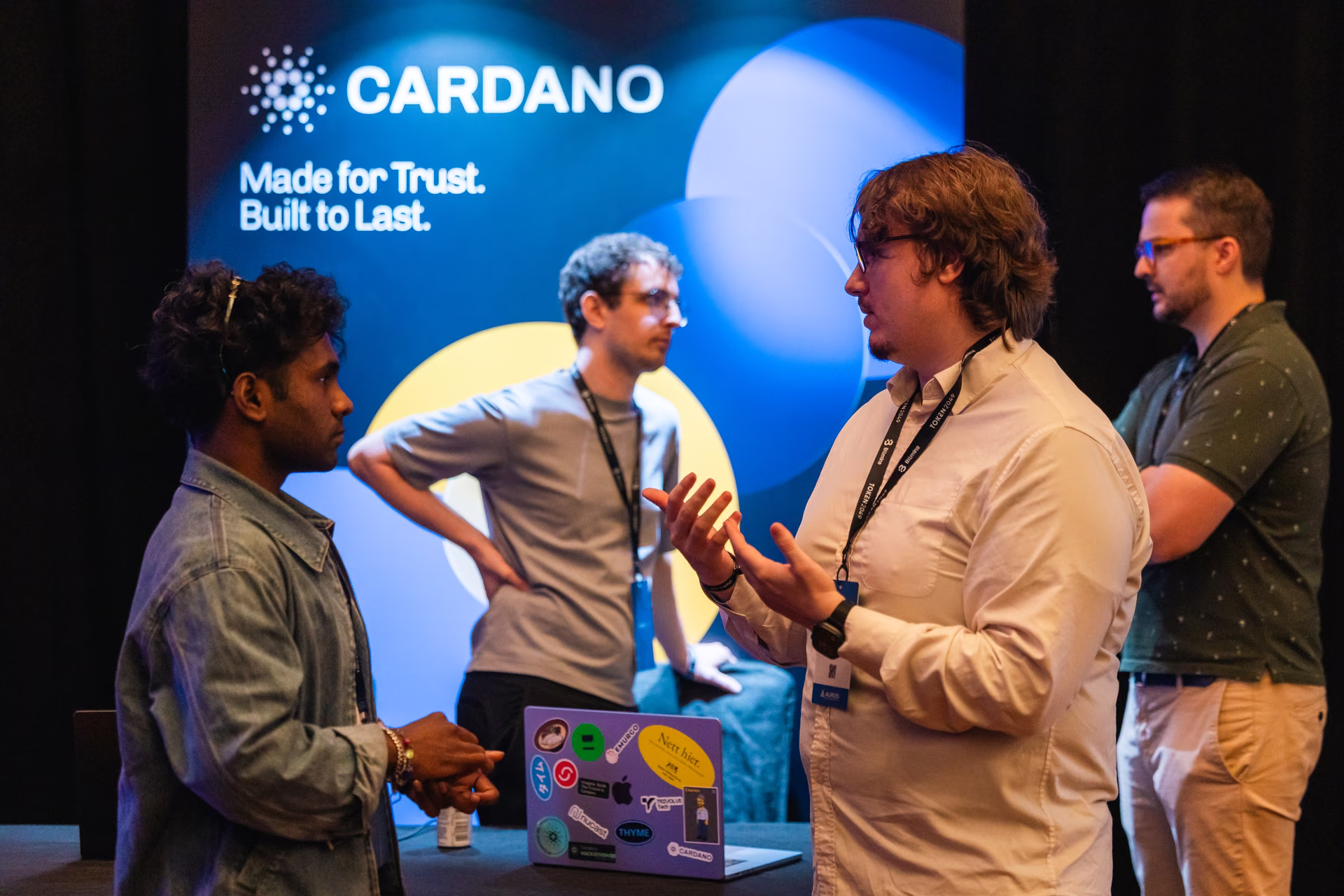 Four men engaged in conversation at a Cardano event booth with a sign displaying 'CARDANO Made for Trust. Built to Last.' and a laptop covered with stickers on the table.