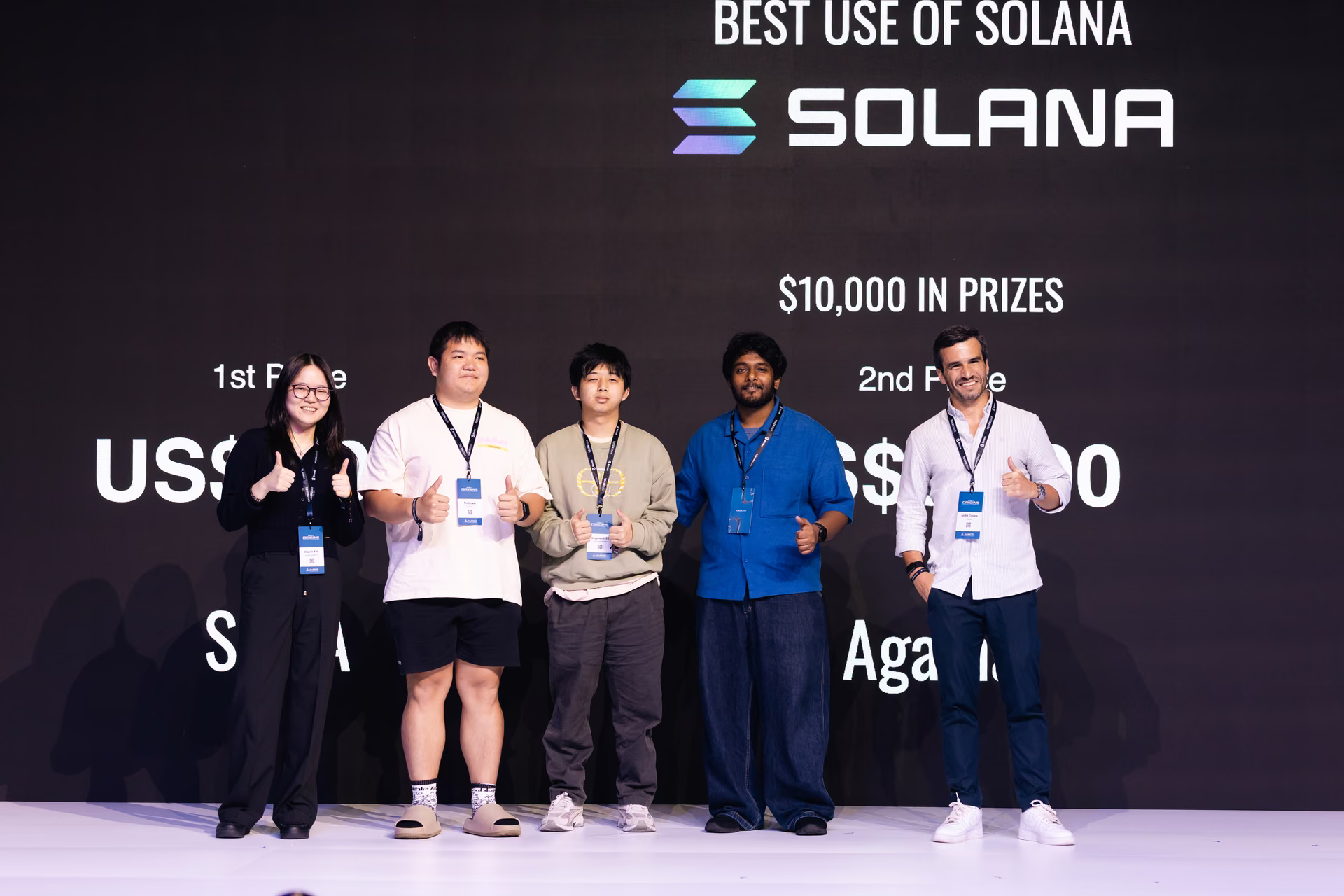 Five people standing on stage in front of a large screen showing Solana logo and prize information, all giving thumbs up gestures.
