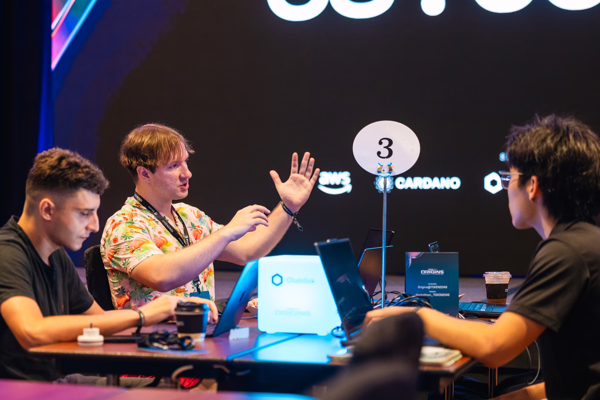 Three young men sit around a table with laptops and coffee, engaged in discussion at a tech event; a table marker labeled '3' is visible and logos for AWS and Cardano appear on a dark background behind them.