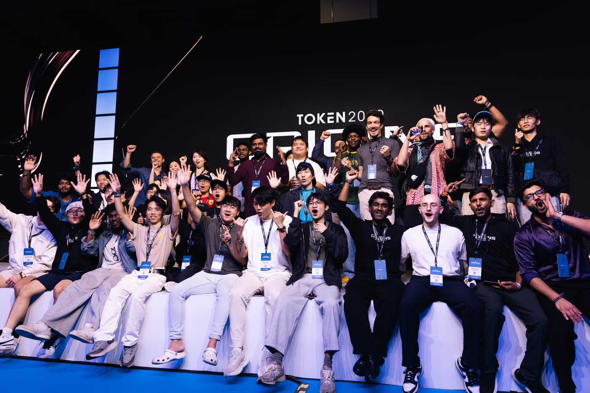Group of diverse young people sitting and standing on a stage with their arms raised, smiling and celebrating at an event with a dark background displaying the partial text 'TOKEN 202'.