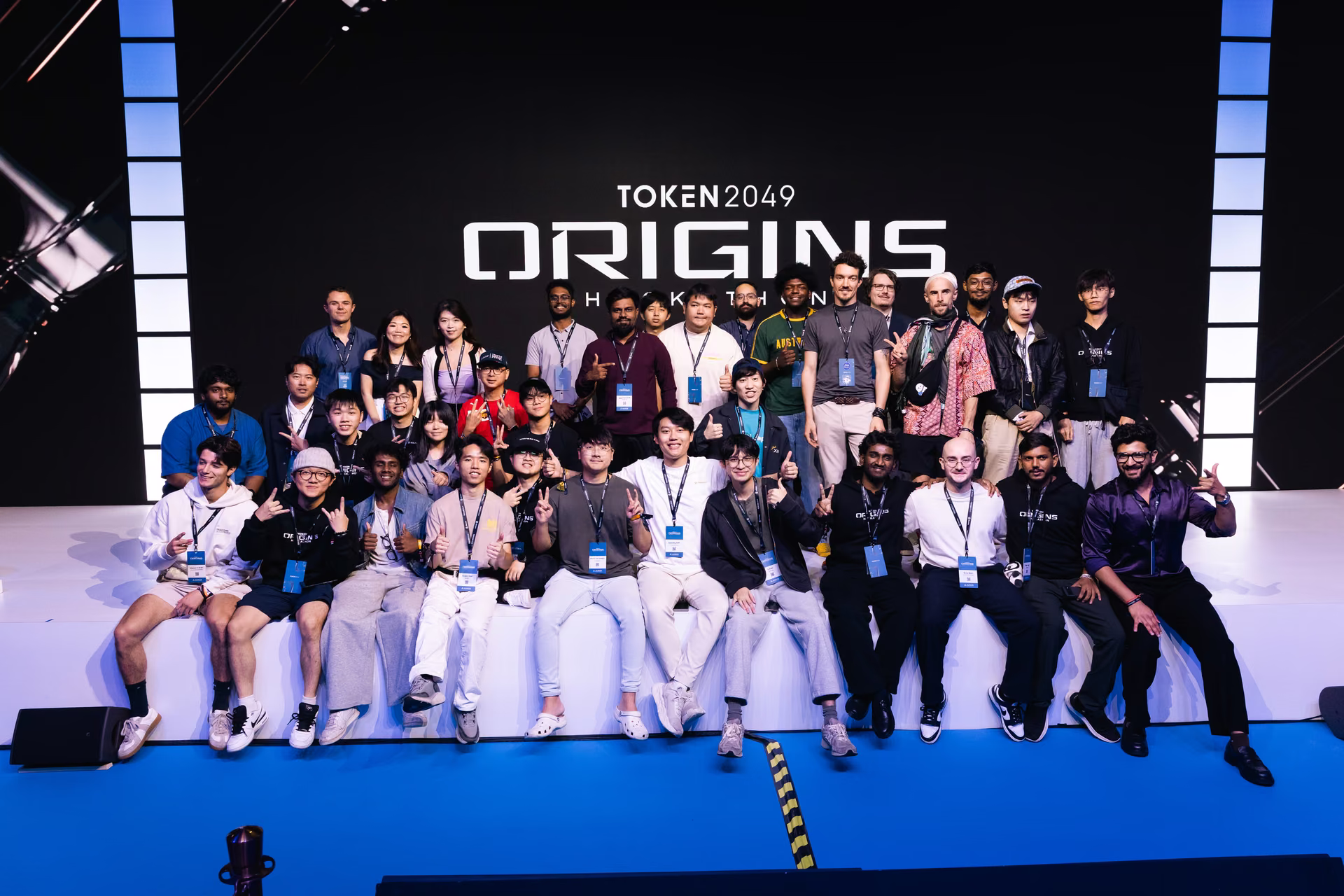 Group of 31 people posing on a stage with a black backdrop displaying the text 'TOKEN 2049 ORIGINS'. The group is smiling and making peace signs, wearing conference badges and casual clothing.