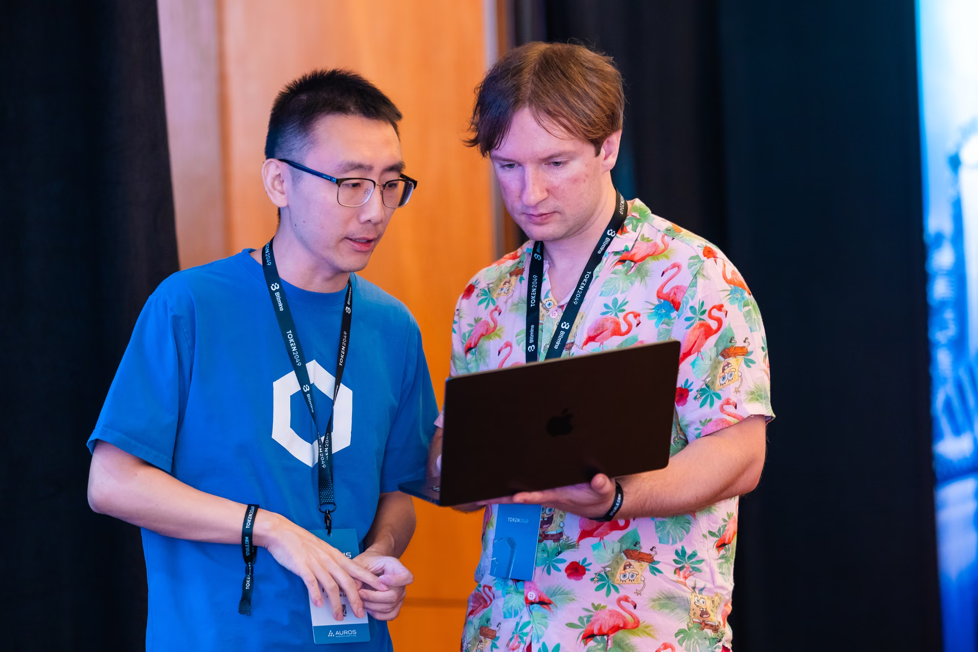 Two men standing side by side reviewing a laptop screen, one wearing glasses and a blue shirt with a geometric design, the other in a pink flamingo-patterned shirt, both wearing event lanyards.