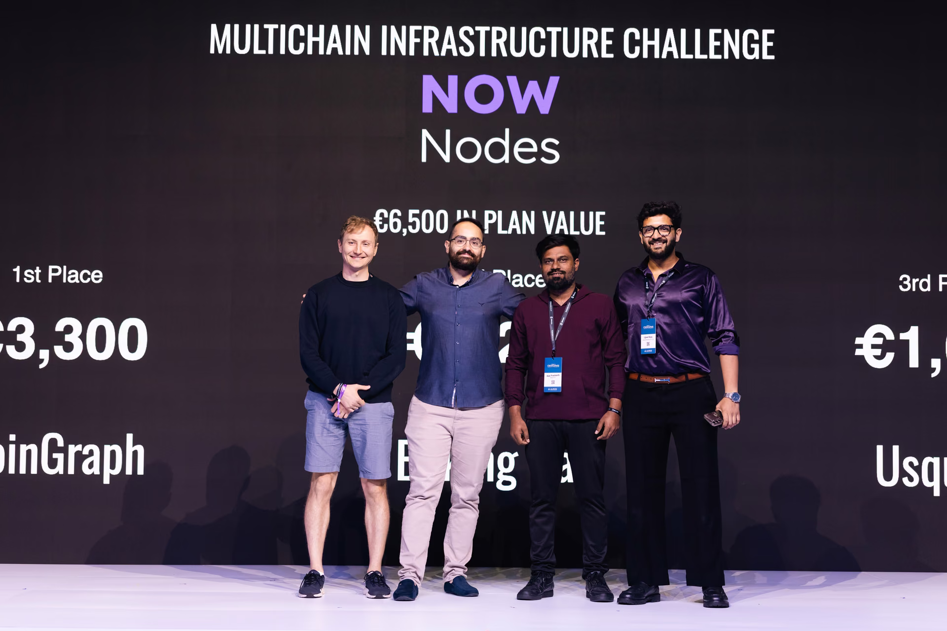 Four men standing on a stage in front of a black backdrop with text about the Multichain Infrastructure Challenge and prize amounts in euros, smiling and posing for a photo.