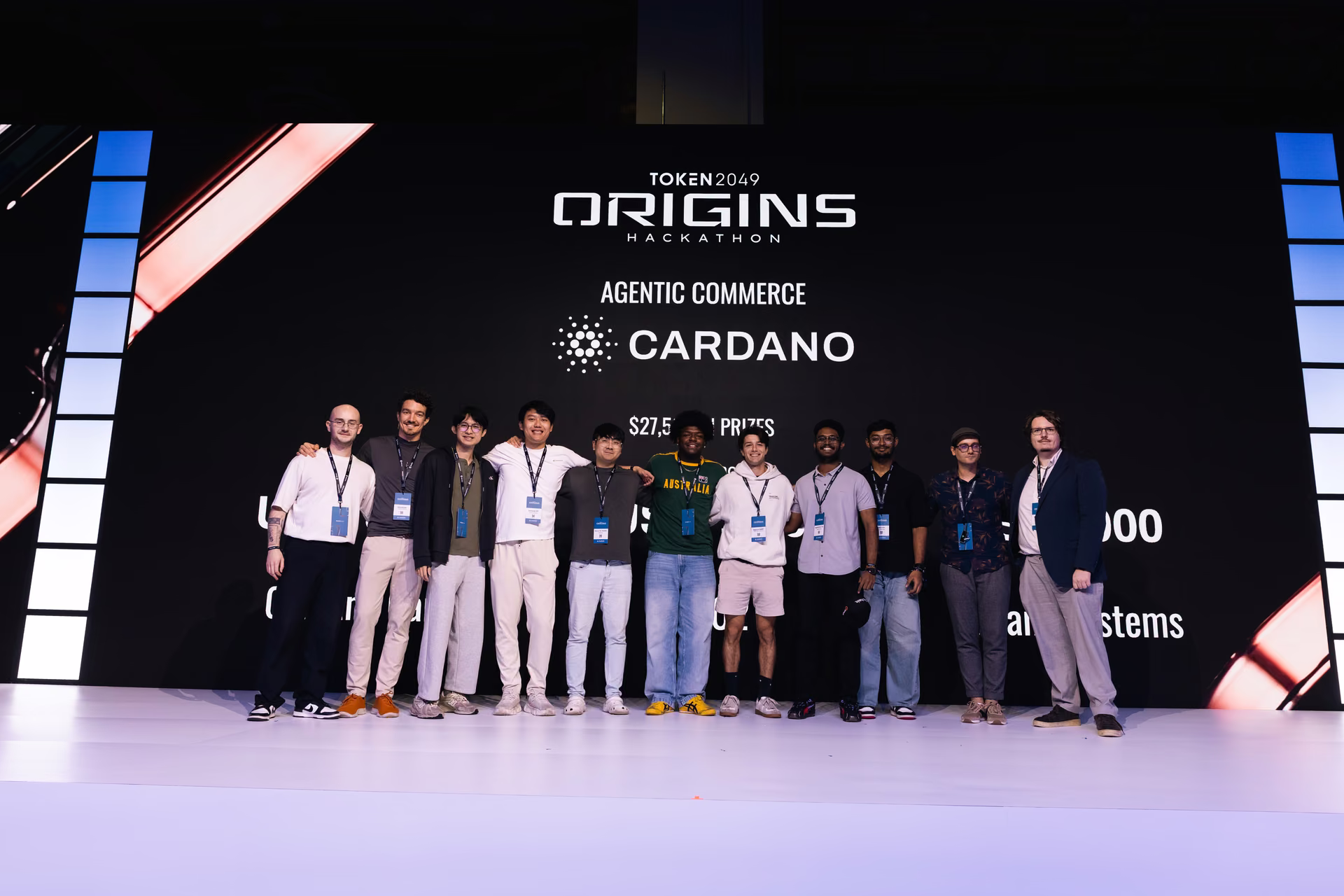 Group of 11 people standing on stage in front of a large screen displaying 'TOKEN2049 ORIGINS HACKATHON', 'Agentic Commerce', 'Cardano', and prize information.