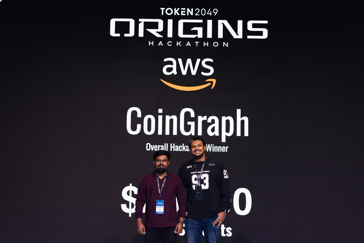 Two men standing and smiling in front of a black backdrop showing 'TOKEN 2049 ORIGINS HACKATHON,' AWS logo, and text announcing CoinGraph as Overall Hackathon Winner with a prize amount partially visible.