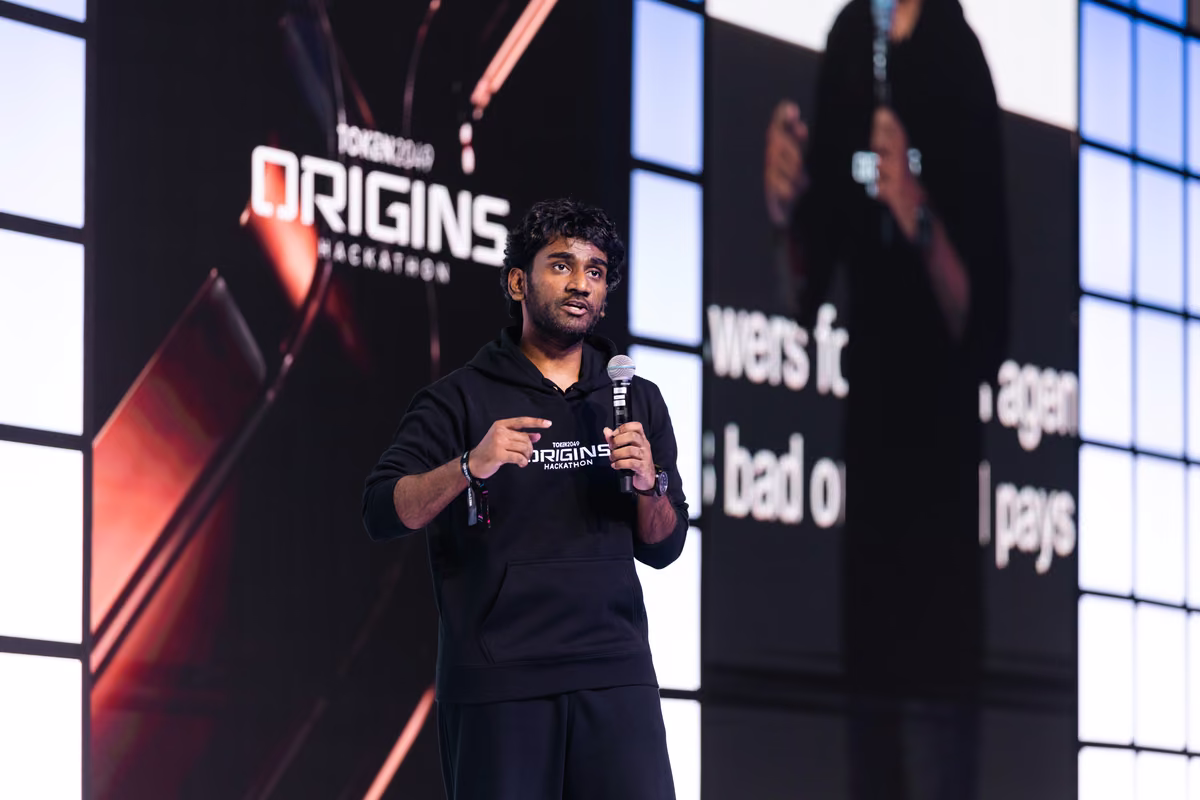 A man wearing a black hoodie with 'TORONTO ORIGINS HACKATHON' printed on it is speaking into a microphone on stage with a large screen behind him showing his image and the event logo.