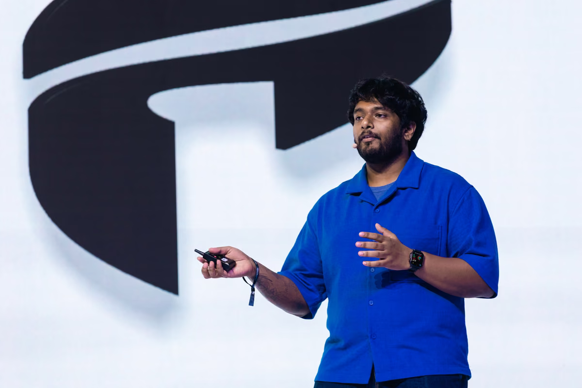 Man in a blue shirt speaking on stage while holding a remote control, with a large abstract black and white background behind him.