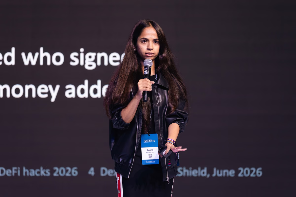 A woman with long dark hair wearing a black jacket and a conference badge stands on stage speaking into a microphone with partial projected text behind her.