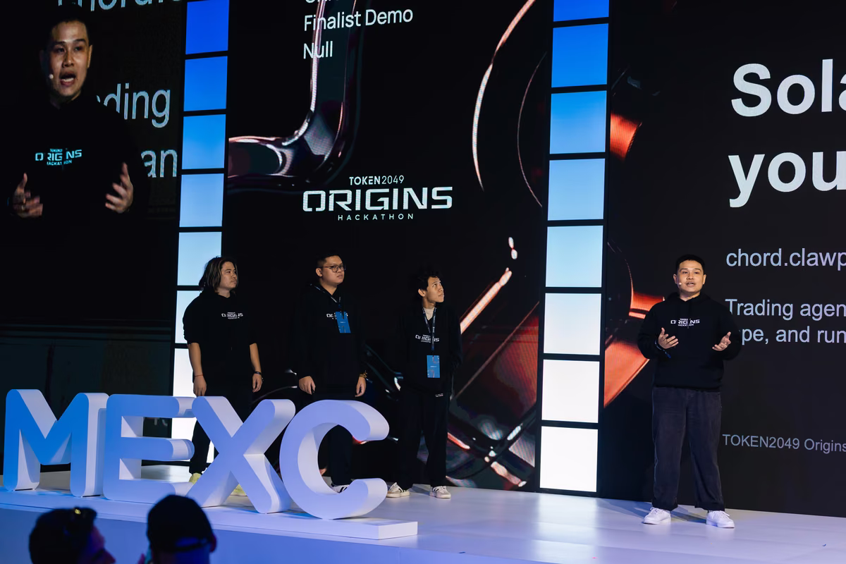 Four people on stage at TOKEN2049 Origins Hackathon, with large lit letters spelling MEXC in front and a presentation screen behind them.