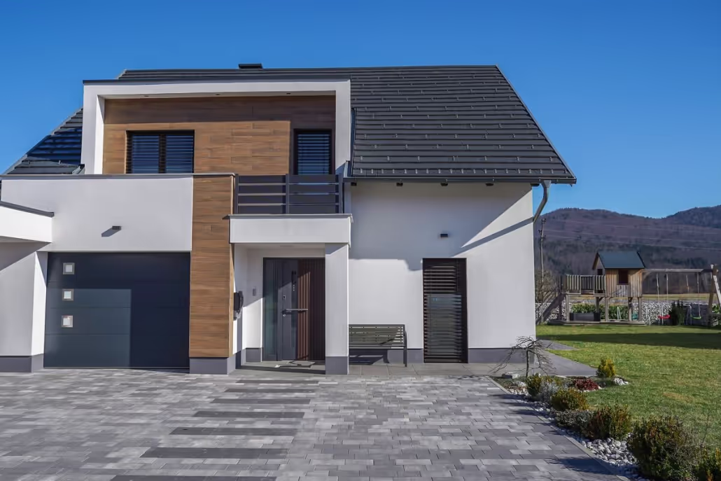 Modernes weißes Haus mit Holzverkleidung, schwarzem Dach, Garage und gepflasterter Einfahrt vor klarem blauem Himmel.