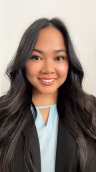 Smiling woman with long black hair wearing a light blue top and black blazer against a plain white background.