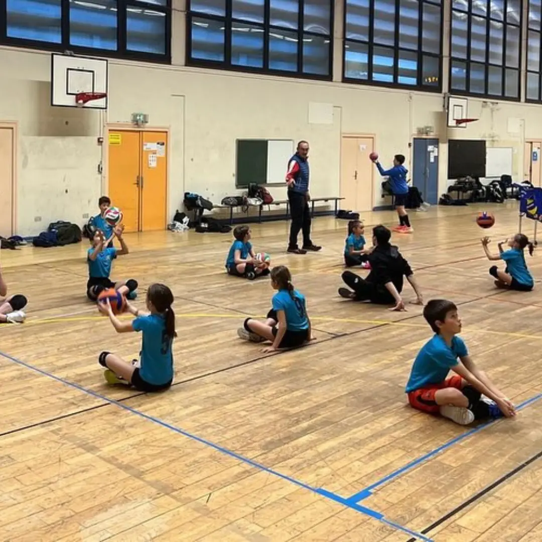 De jeunes joueurs du club Lyon PESD volley, assis au sol, pratiquent un exercice de réception en passe et en manchette. Cette scène met en avant leur apprentissage et la transmission des fondamentaux du volley dans une ambiance conviviale et studieuse.
