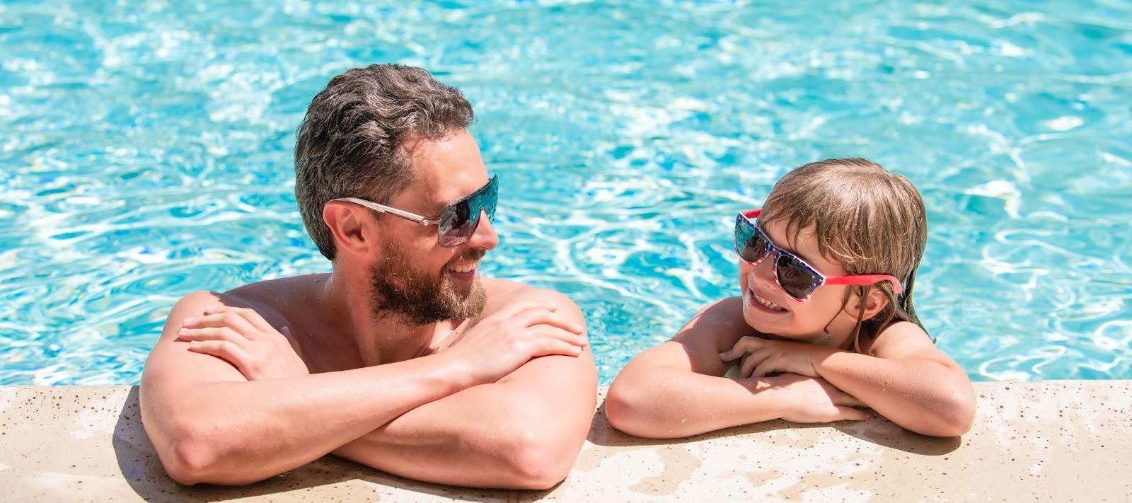 Father and child in sunglasses smiling at each other while resting at the edge of a pool on a sunny day