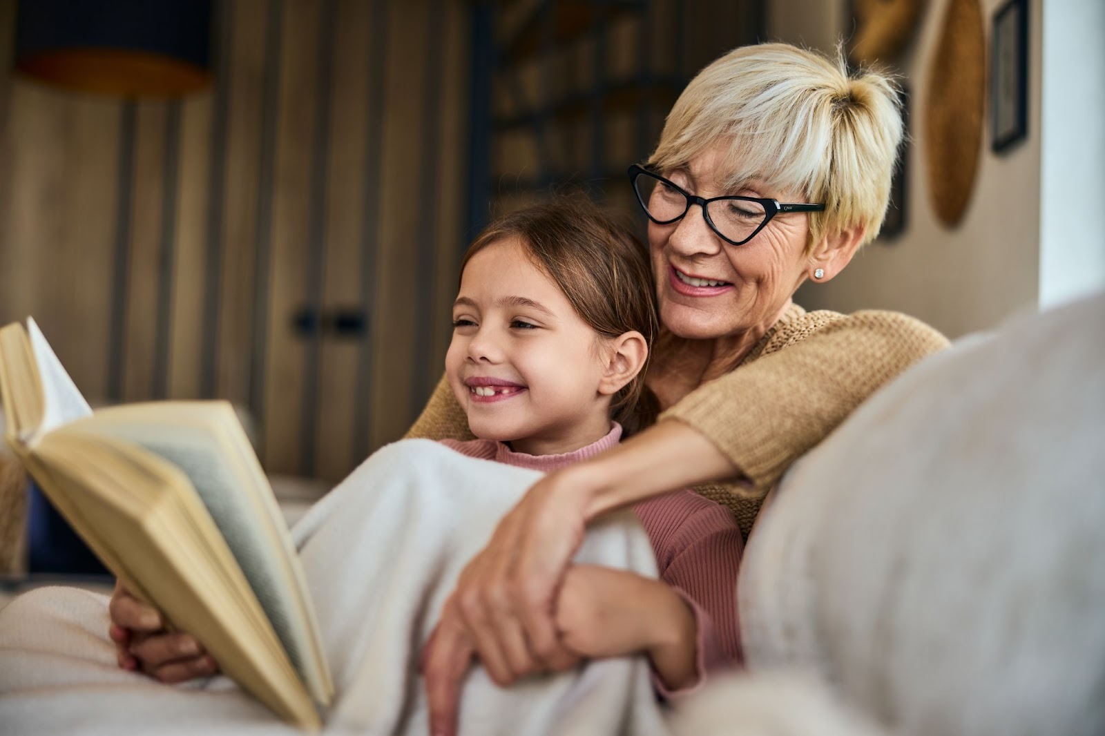 Smiling grandma and her grandchild reading a book at home while sitting on the couch