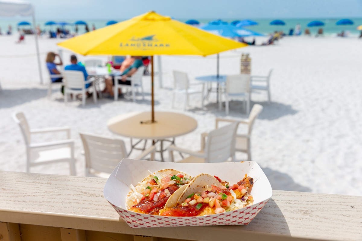  An order of fish tacos sitting on a wooden ledge with beachfront seating in the background.
