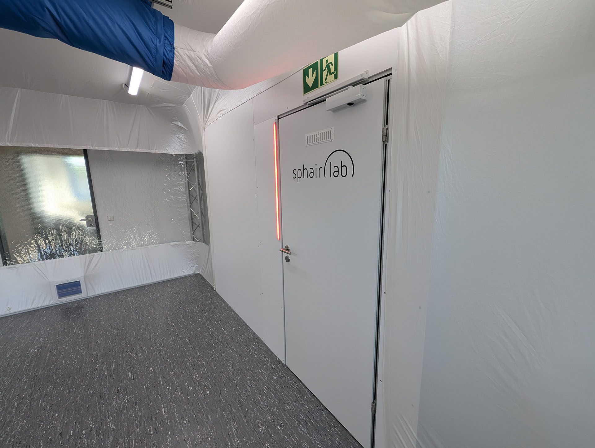 Entrance area of a cleanroom with a white door and sphairlab logo. A green and white emergency exit sign is mounted above the door, and a blue air duct runs along the ceiling.