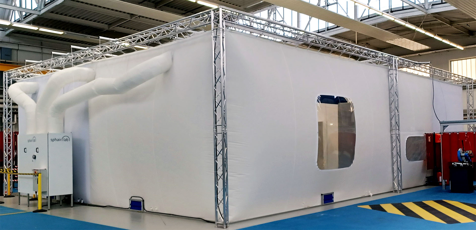 Large modular cleanroom tent from sphairlab in an industrial hall, supported by an aluminum frame structure. Several white air hoses connect to the cleanroom systems, and viewing windows are integrated into the side panels.
