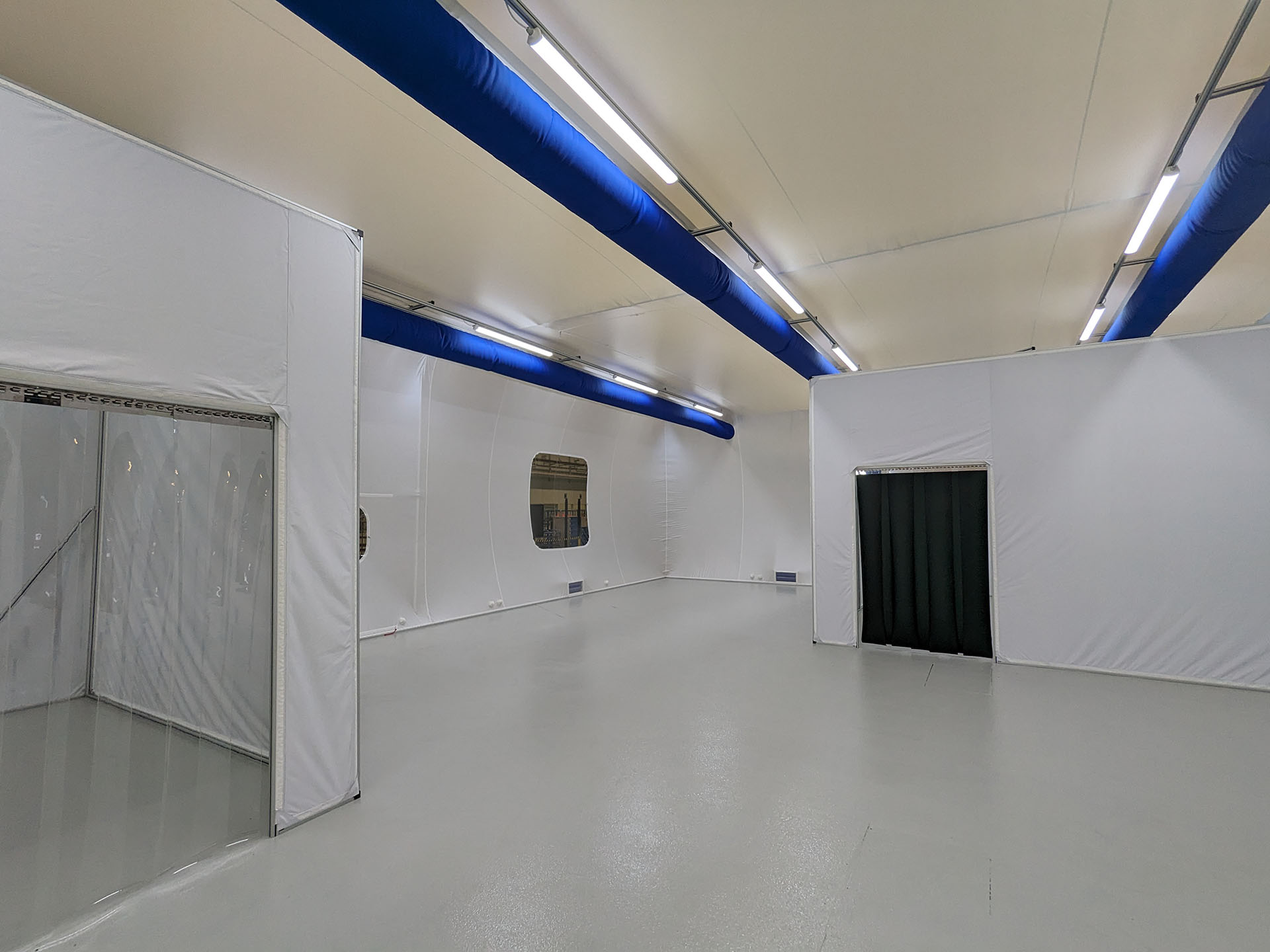 Interior of a modular cleanroom with white walls, smooth industrial flooring, and blue air ducts along the ceiling. Viewing windows and flexible entrance curtains separate individual work areas.