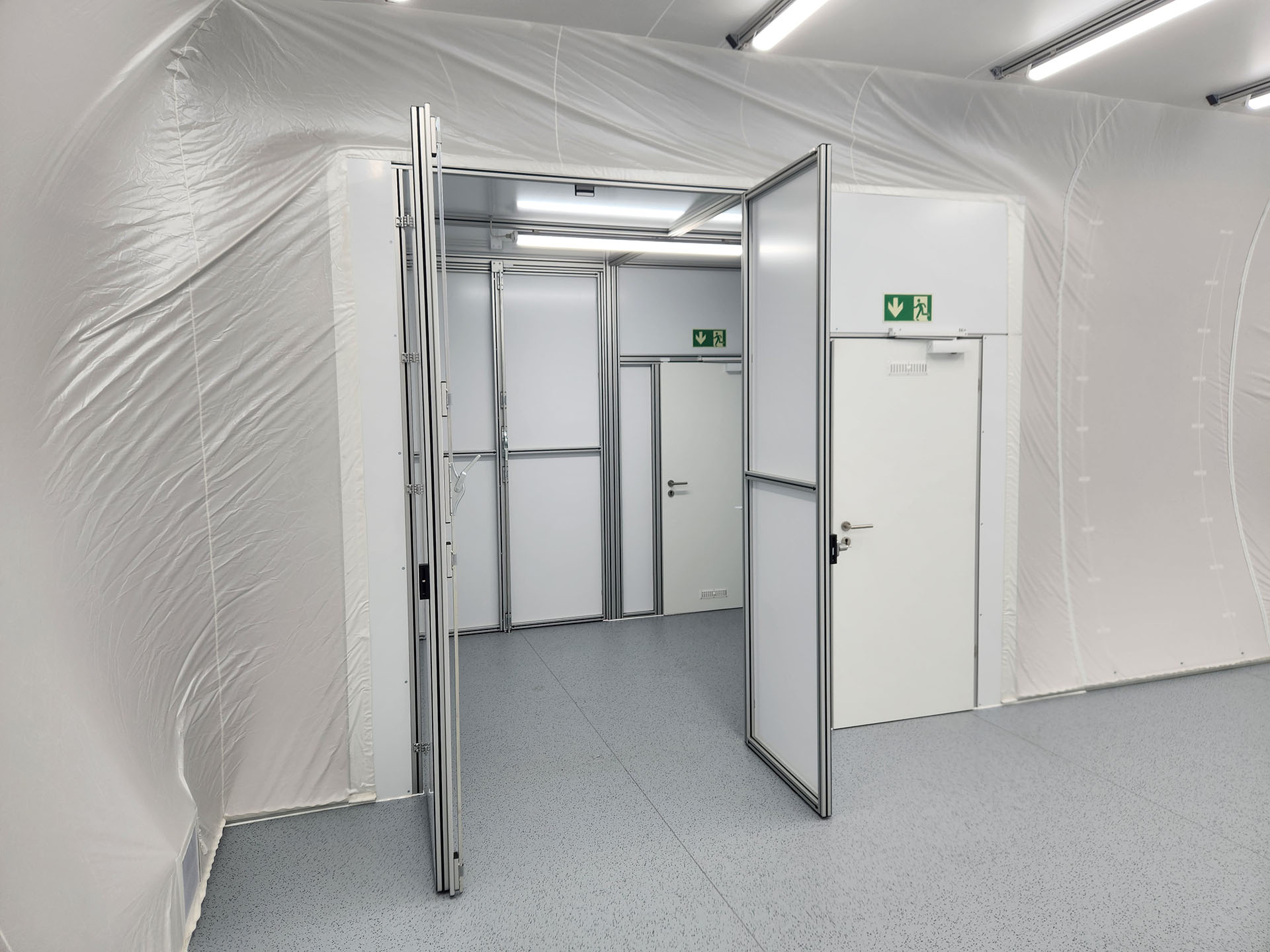 Innenansicht eines modularen Reinraums mit weißen Wänden und leicht zu reinigendem Boden. Mehrere Türen und Aluminiumrahmen trennen Schleusen- und Arbeitsbereiche, darüber gut sichtbar grün-weiße Notausgangsschilder.
