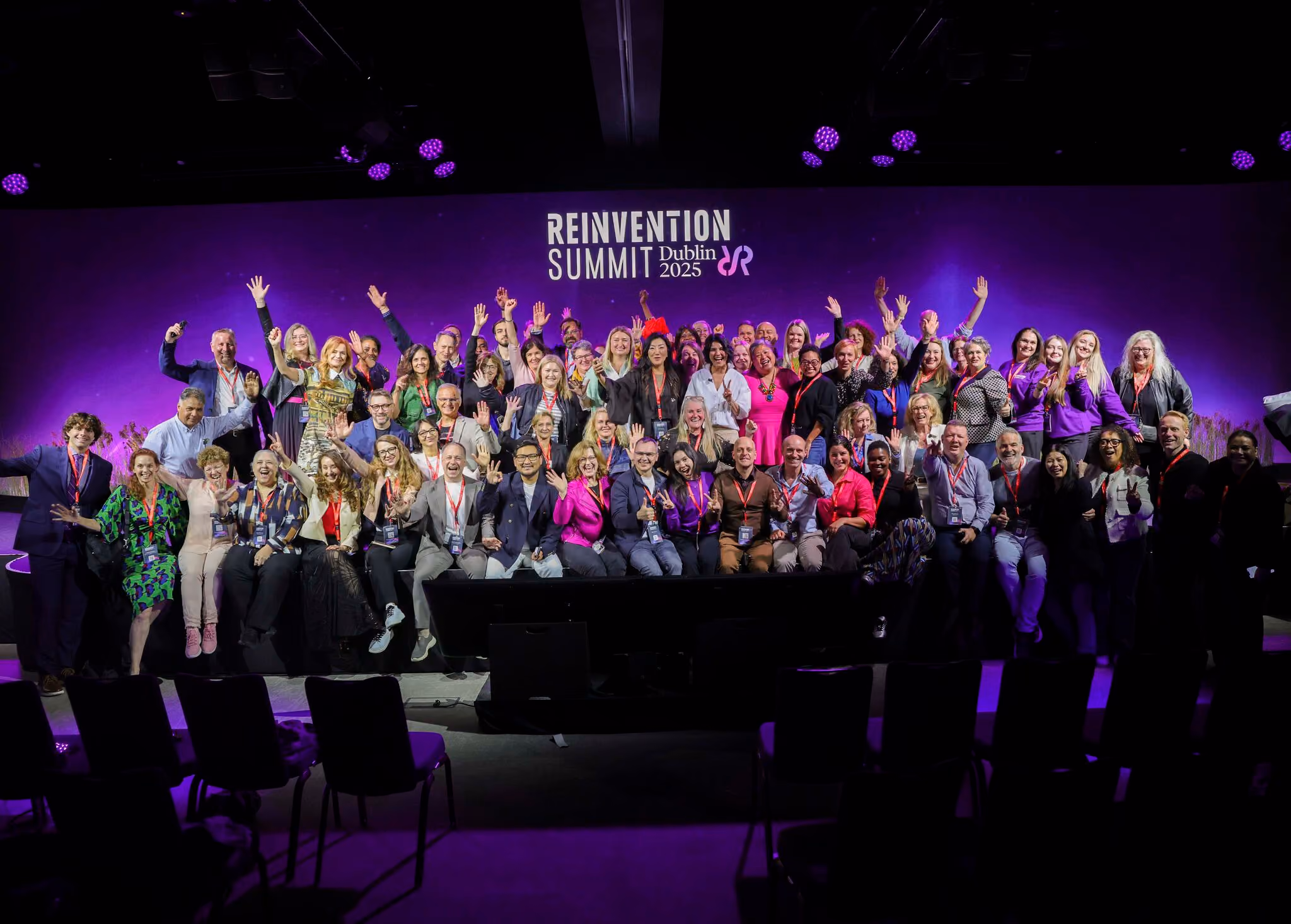 Group of diverse people posing and waving in front of a purple backdrop at Reinvention Summit Dublin 2025.