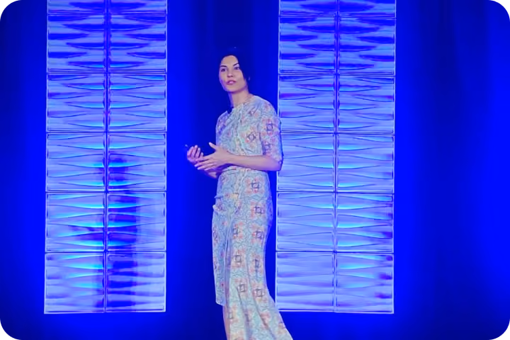 Woman in a floral dress speaking on stage with blue lit wall panels behind her.